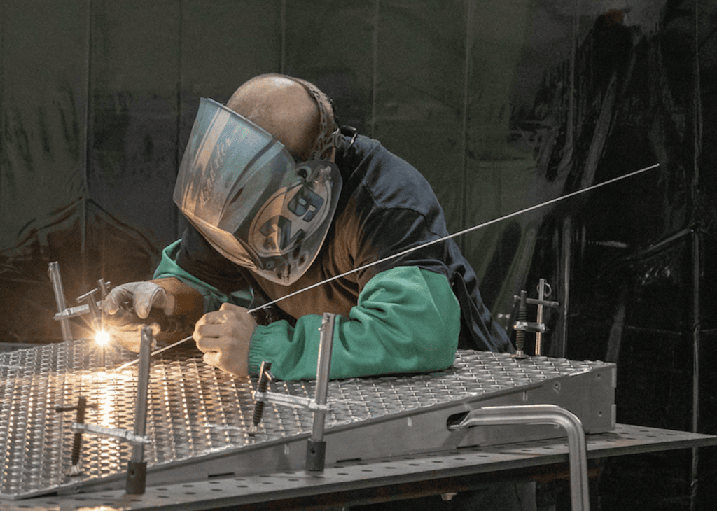 Man in welding mask holding tools and working on a metal fabrication project.